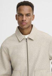 Beige uldblandet jakke med krave, frontlynlås, to lommer og en blød tekstur. Enkel, minimalistisk design.