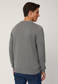 Maglione grigio a maglia con colletto rotondo, polsini e orlo a costine. Il tessuto sembra morbido e di spessore medio. Indossato sopra jeans blu.