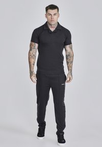 Polo negro de punto con cuello, combinado con pantalones jogger oscuros y ajustados que presentan un acento de logo. Llevado con zapatillas negras.