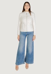 Giacca bianca trapuntata con colletto alto e zip frontale, abbinata a jeans blu a gamba larga e scarpe nude. Sfondo neutro.