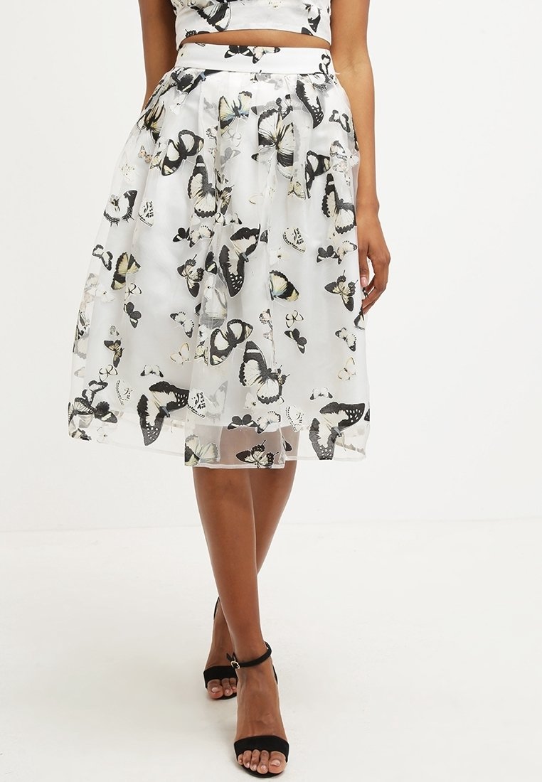Femme portant une jupe midi blanche à motif papillons, un top assorti et des talons noirs ouverts avec bride à la cheville, debout sur un sol blanc.
