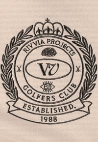Bež tkanina s crnim izvezenim okruglim logotipom na kojem se nalazi tekst "Rivvia Projects Golfers Club" i godina "1988", okružen lovorovim grančicama.