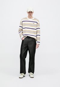 Majica dugih rukava s prugama u bež boji s ljubičastim i bijelim prugama, u kombinaciji s crnim hlačama i bijelim tenisicama. Kapa za bejzbol svijetle boje.