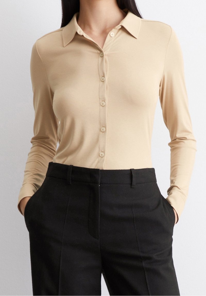 Femme portant une chemise beige à manches longues boutonnée rentrée dans un pantalon noir taille haute, les mains dans les poches.