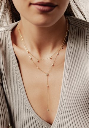 Goldfarbene Halskette mit einer Schicht, die kleine Herz- und Diamantakzente aufweist, mit einer zarten Kette und einem V-förmigen Anhängerdesign. Beigefarbenes, strukturiertes Oberteil sichtbar.