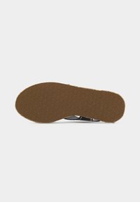 Semelle d'une sandale noire avec une semelle inférieure en caoutchouc marron texturée et de petits clous dorés visibles sur la partie supérieure.