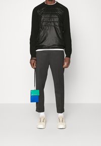 Czarny sweter z pikowanym, błyszczącym przodem, zestawiony z szarymi spodniami. Białe sneakersy i wielokolorowy portfel zawieszony w dłoni.