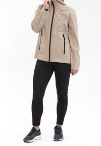 Veste d'extérieur beige avec une capuche, fermeture éclair à l'avant et deux poches latérales, portée sur des leggings noirs et des chaussures de sport grises. Texture de tissu lisse.