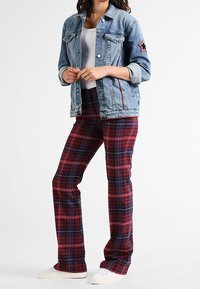 Jeansjacka med röd sömnad och stjärnpatch, bärs över en vit tanktopp, kombinerad med röd- och blårutiga utsvängda byxor och vita sneakers.