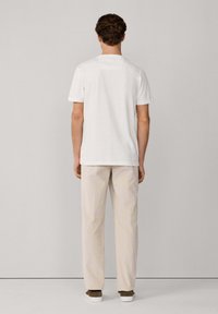 Camisa blanca de manga corta con dobladillo recto, combinada con pantalones de color beige claro y zapatos oscuros, vista desde atrás.