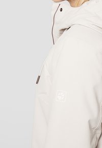 Personne portant une veste à capuche beige clair avec une fermeture éclair et des cordons, comportant un petit logo en forme d'empreinte de patte sur le haut de la manche.