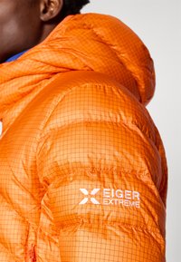 Casaco puffer laranja com um padrão quadriculado e capuz. As características incluem um logótipo na manga e uma superfície de tecido texturizada.