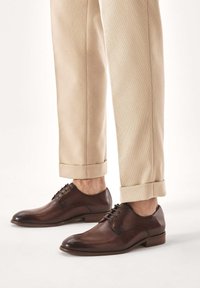 Pantofi eleganți din piele maro, cu finisaj lustruit, vârf rotunjit și detaliu cu șireturi. Asortați cu pantaloni bej deschis, cu tiv răsucit.