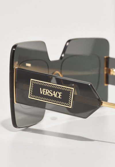 Lunettes de soleil oversize noires avec des verres sombres, dotées d'une charnière à accents dorés et d'une monture rectangulaire marquée d'un logo "Versace" sur le côté.