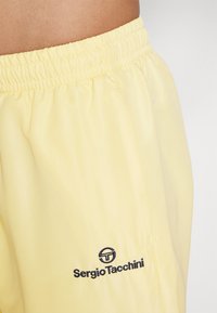 Lysegule treningsshorts laget av glatt, pustende stoff med elastisk midje og en mørkeblå logo på venstre side.