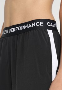 Lähikuva vyötaresta, jossa on päällä mustat Calvin Klein Performance -shortsit, joissa on valkoinen sivuraita ja joustava brändinauha vyötäröllä.