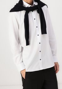 Weißes Hemd mit langen Ärmeln, schwarzen Knöpfen und einem schwarzen Pullover, der über die Schultern gelegt ist. Das Outfit wird mit schwarzen Hosen kombiniert.