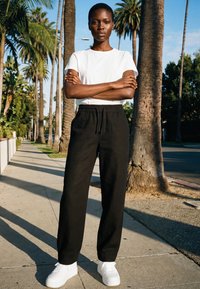 Frau steht mit verschränkten Armen auf einem sonnigen Bürgersteig, der von hohen Palmen gesäumt ist, trägt ein weißes T-Shirt, schwarze Hosen und weiße Turnschuhe.