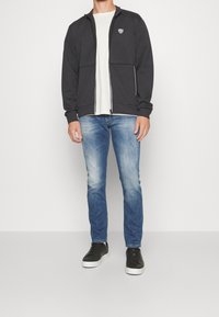 Tmavě šedá mikina na zip s dvěma předními kapsami, kombinovaná s světle modrými džíny a černými teniskami. Mikina má na hrudi logo.