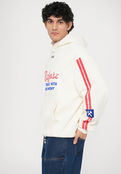 Witte capuchontrui met rode strepen op de mouwen, grafische tekst in roze en blauw, voorzak en blauwe patch met een embleem. Denim onderkleding.