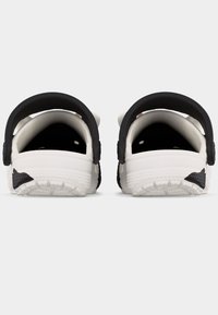 Ζευγάρι λευκών και μαύρων παπουτσιών τύπου «κλογ» χωρίς κορδόνια, που φαίνονται από πίσω, με ρυθμιζόμενα λουράκια στη φτέρνα και υφασμένα πέλματα σε φωτεινό φόντο.