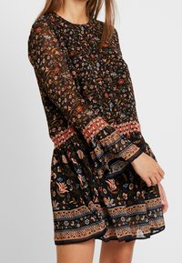 Mujer vestida con un vestido floral negro transparente con flores rojas y azules, puños y dobladillo de patrón geométrico, de pie contra un fondo liso.