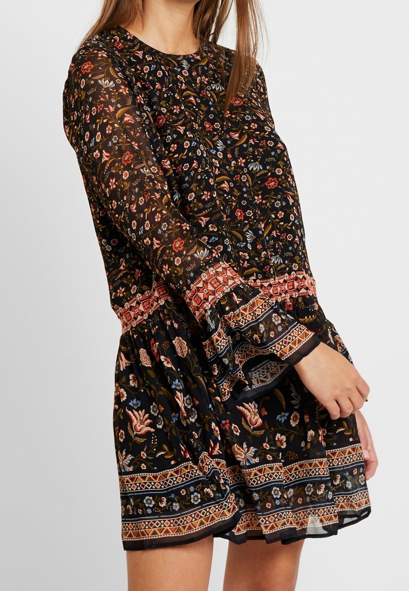 Mujer vestida con un vestido floral negro transparente con flores rojas y azules, puños y dobladillo de patrón geométrico, de pie contra un fondo liso.