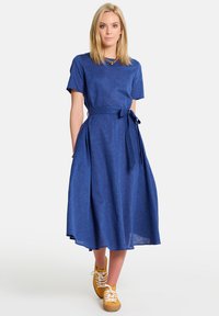 Sinine linane kleit lühikeste varrukatega, vöökoht ja laienev seelik, koos kollaste spordijalatsitega. Sile tekstuur ja minimalistlik disain.