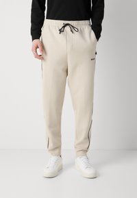 Beige sweatpants med sorte sidelæder, elastisk talje og en snøre. Logoet "BOSS" trykt på siden. Sammen med hvide sneakers.