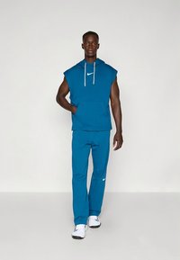 Hoodie azul sem mangas com cordões brancos e logótipo, combinada com calças azuis a condizer. Tecido suave, ajuste descontraído e design até ao tornozelo.