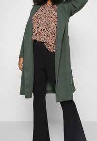 Cardigan largo verde oliva hecho de tela suave, con frente abierto y mangas anchas, combinado con una blusa de estampado floral y pantalones de campana negros.