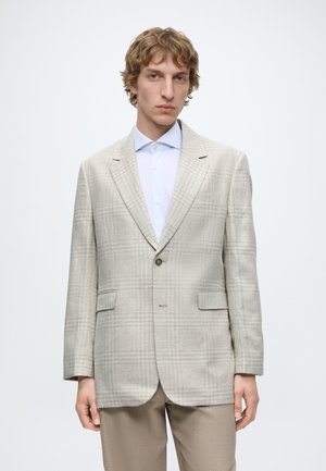 Jeune homme aux cheveux blonds ondulés portant un blazer à carreaux beige clair, une chemise blanche et un pantalon beige, debout devant un fond blanc.