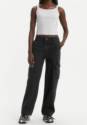 Persona joven con top corto blanco, pantalones cargo negros de pierna ancha y zapatillas deportivas negras y grises, de pie con una mano en el bolsillo.