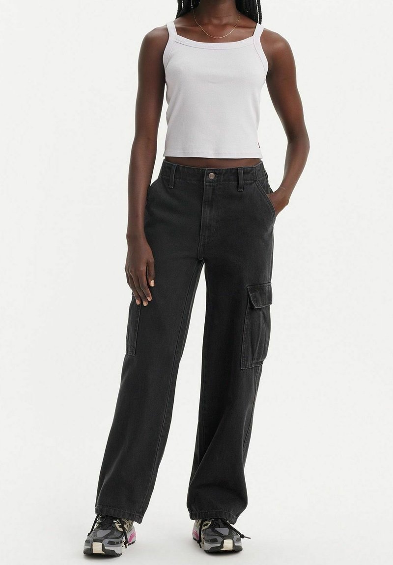 Jeune personne portant un débardeur court blanc, un pantalon cargo noir à jambes larges et des chaussures de sport noires et grises, debout avec une main dans la poche.