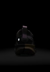 Czarno-brązowe sneakersy widziane z tyłu, z materiałową pętelką i białym okrągłym detalem na pięcie, w słabym oświetleniu.