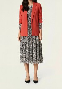 Blazer rosso su un vestito floreale bianco e nero con gonna a strati. Il vestito ha piccoli dettagli rossi e maniche lunghe e trasparenti. Tacchi a punta neri.