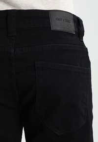 Pantalones vaqueros de mezclilla negra con una textura suave, que cuentan con una etiqueta parche en la cintura y un bolsillo en la parte trasera del lado derecho.