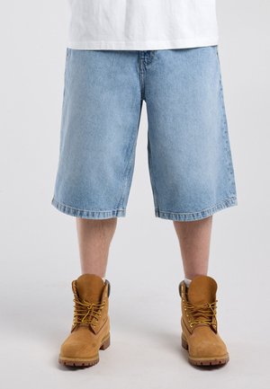 Lichtblauwe denim knielange korte broek gedragen met beige veterwerklaarzen en witte sokken, getoond van taille tot voeten op een neutrale achtergrond.