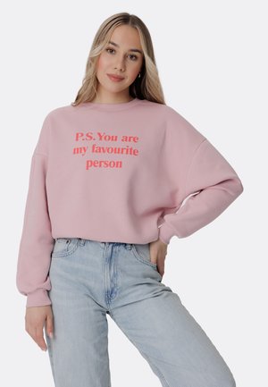 Sweatshirt rose avec le texte rouge "P.S. Tu es ma personne préférée," présentant une coupe décontractée et des poignets côtelés. Associé à un jean bleu clair.