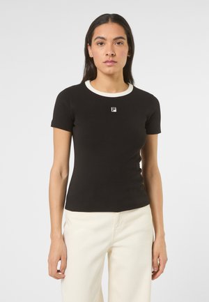 Donna che indossa una maglietta nera aderente con colletto bianco e piccolo logo, abbinata a pantaloni crema a vita alta, in piedi davanti a uno sfondo bianco.