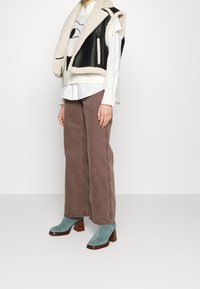 Schwarze und cremefarbene Lammfellweste über einer weißen Bluse, kombiniert mit braunen weiten Hosen und türkisfarbenen Stiefeletten mit Holzabsatz.