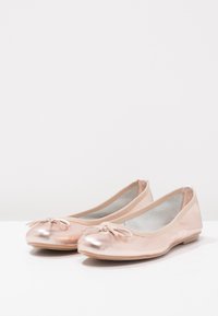 Bailarinas metálicas de color rosa con punta redondeada y un pequeño lazo en la parte delantera. Fabricadas en material sintético suave.
