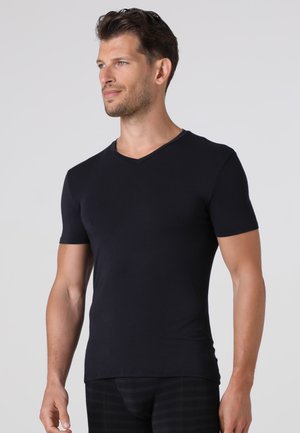 Uomo in piedi con postura rilassata che indossa una maglietta nera aderente a maniche corte con scollo a V e pantaloni neri davanti a uno sfondo grigio chiaro.