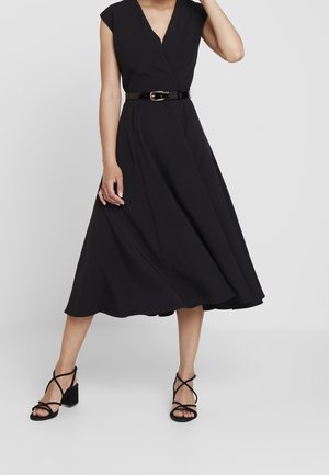 Femme portant une robe midi noire sans manches avec un col en V, une ceinture noire brillante et des sandales à talons blocs noirs à brides, debout devant un fond gris.