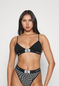 Čierny bralet s tenkými ramienkami a trojuholníkovými pohárikmi, s šedým pásom s logom Calvin Klein. Zodpovedajúce čierne a biele vzorované tangá.