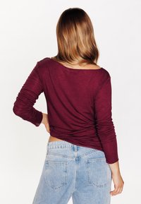 Top bordeaux a maniche lunghe con scollatura ampia e texture arricciata sulla schiena, abbinato a jeans in denim azzurro chiaro a vita alta.