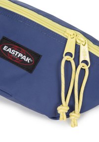 Blå tygpåse med en gul dragkedja med rundade draghandtag och en svart Eastpak-logotyp. Texturerad yta med hållbara sömmar.