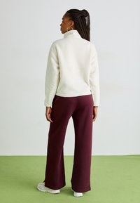 Vit fleece-tröja med hög krage, parat med mörk burgundy vida byxor och vita sneakers. Flätat hår, neutral bakgrund.