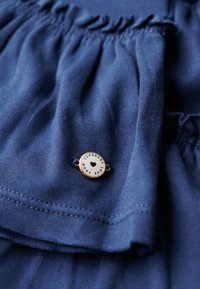 Tela azul marino con detalle fruncido, con un botón circular de tono dorado. El botón tiene la inscripción "SUPERDRY" y un diseño de corazón. Textura suave.