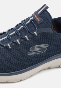 Baskets en maille bleu marine avec lacets élastiques et accents gris. Présentent un dessus texturé et une semelle extérieure en caoutchouc blanc avec un logo distinct.